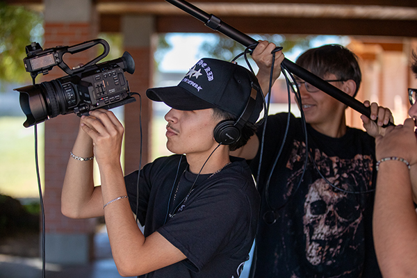 A teen boy holds a video camera while a teen boy stands behind him with a boom mic
