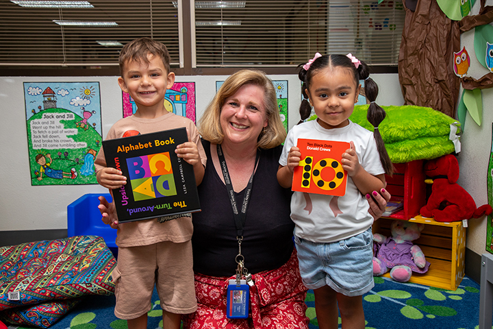 A woman, middle, smiles with a boy and a girl holding books in a pre-k classroom