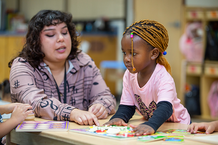 A woman in a purple shirt works with a little girl in a pink shirt on a classroom game