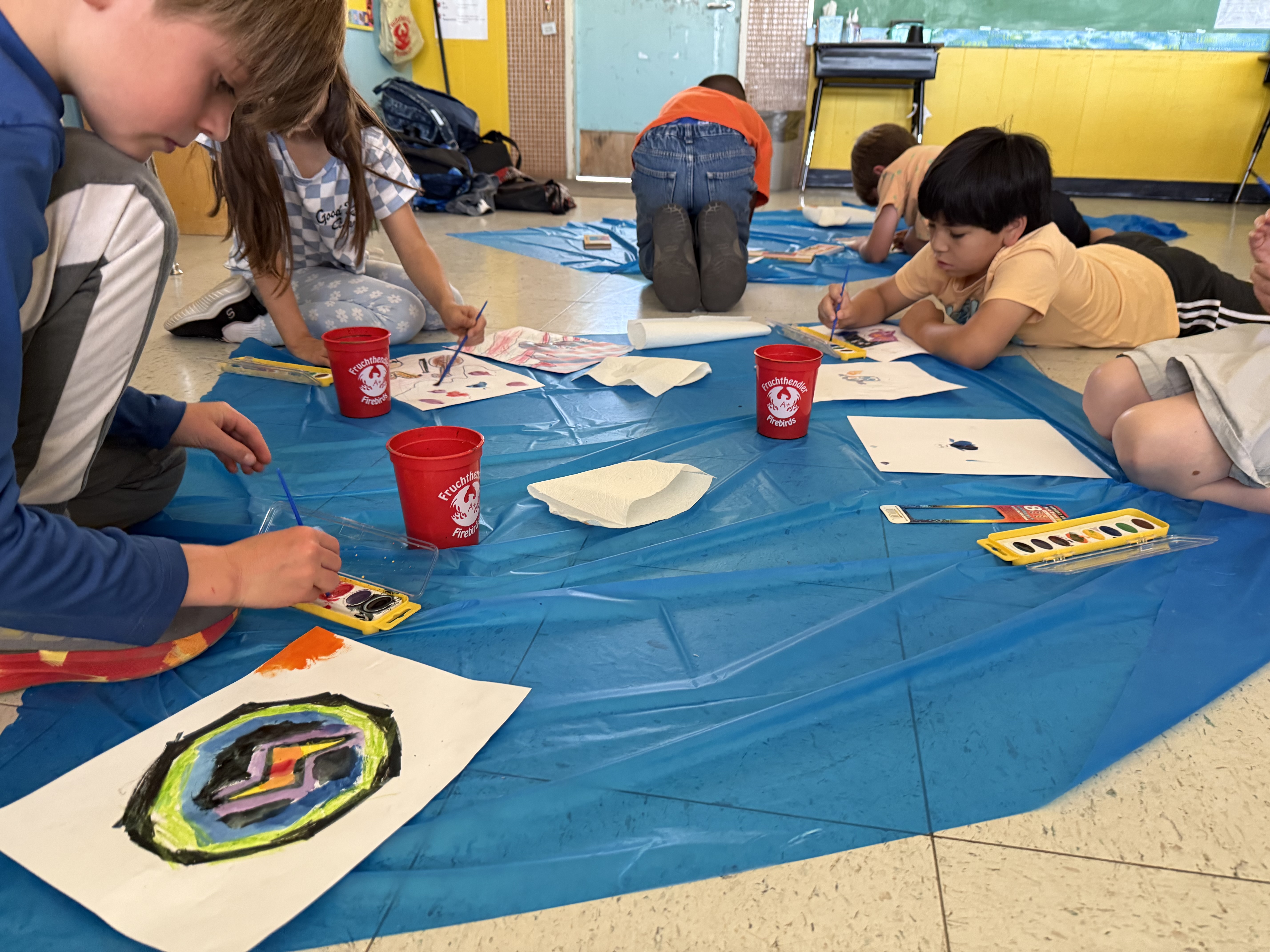 Students painting with watercolors while laying on the ground over a blue tarp to keep the paint off the floor.