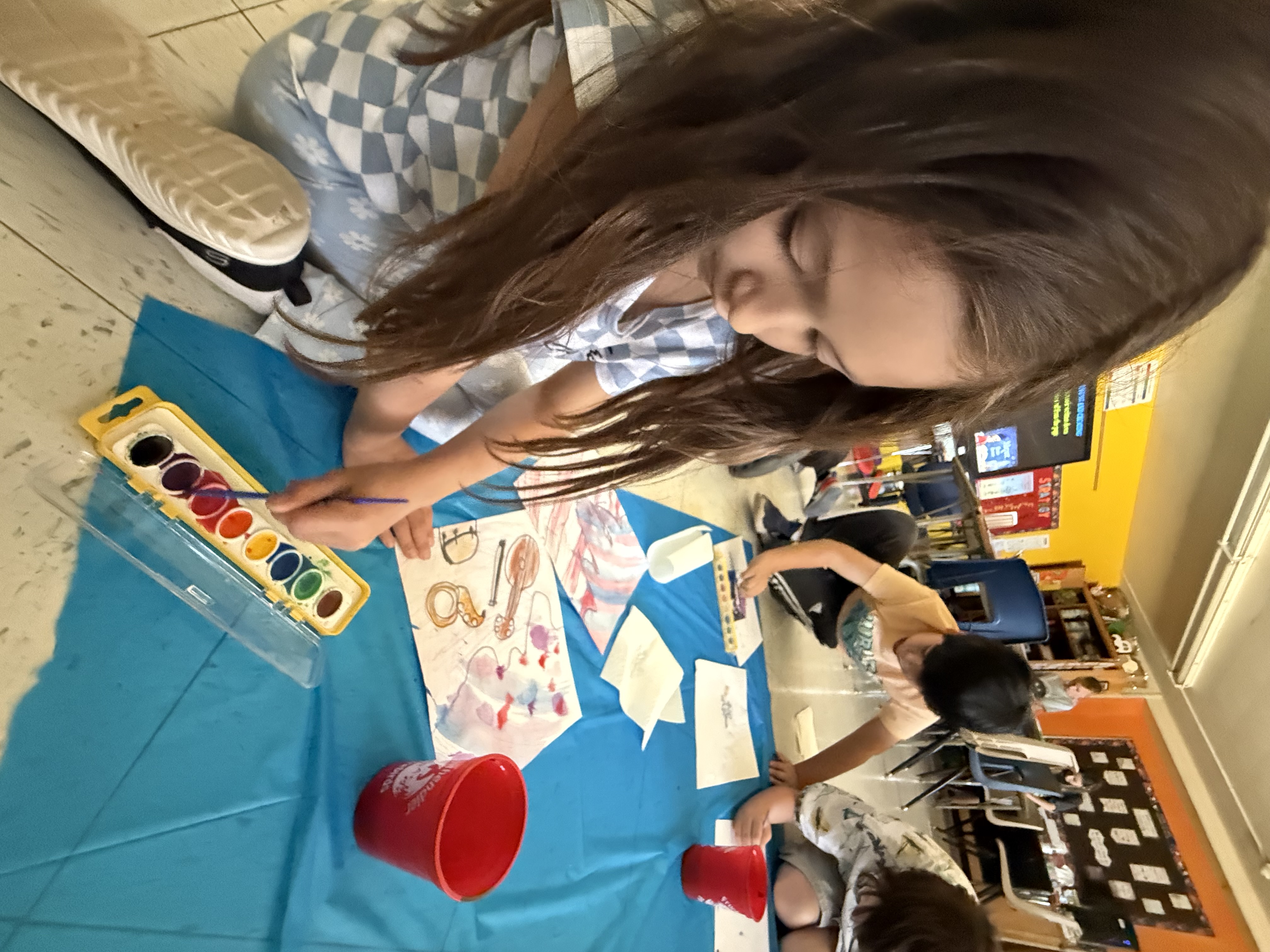 Students painting with watercolors while laying on the ground over a blue tarp to keep the paint off the floor.
