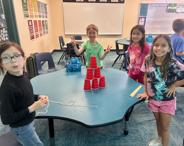 This picture shows students participating in a team game building a cup tower as part of our Olympics of the Mind unit, engaging with one of Gardner’s multiple intelligences.
