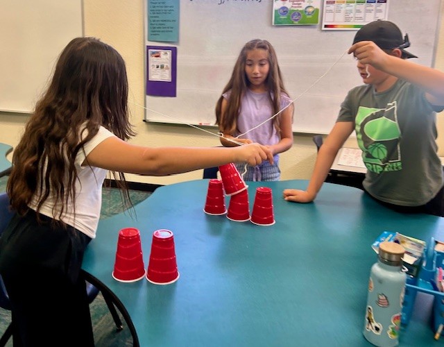 This picture shows students participating in a team game building a cup tower as part of our Olympics of the Mind unit, engaging with one of Gardner’s multiple intelligences.