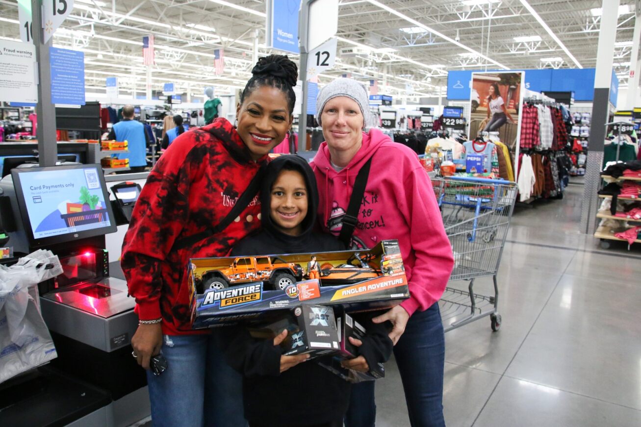 A boy smiles with his new truck and boat toy, alongside two adults.