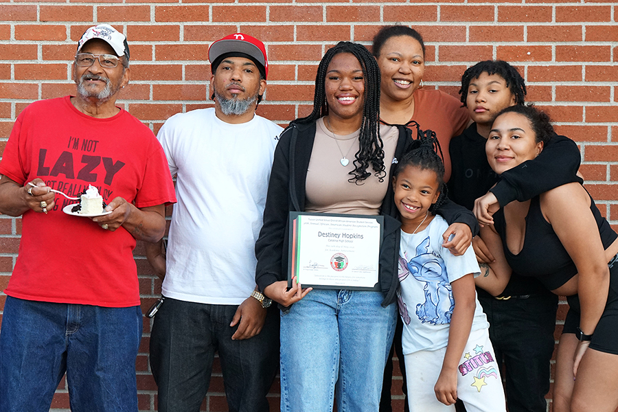 A teen girl holding an award certificate smiles, surrounded by several family members