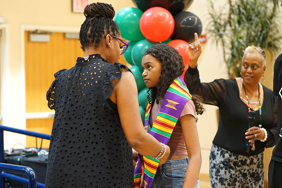 A woman in a black lacy top places a colorful sash around a girl's shoulders