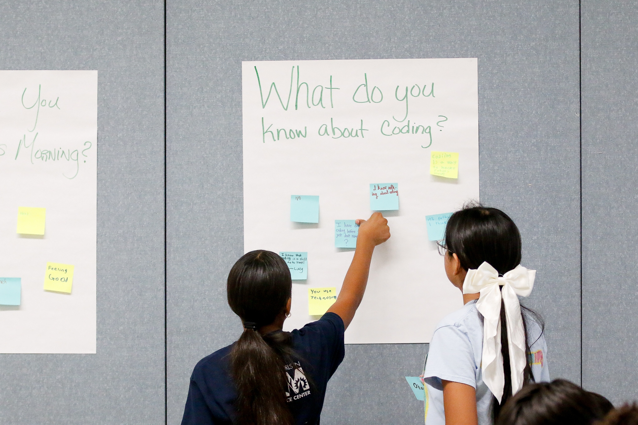 Two girls stick post-it notes on a poster reading What do you know about coding?