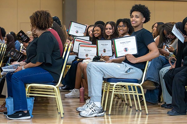 Students sitting in the audience proudly hold up their award certificates