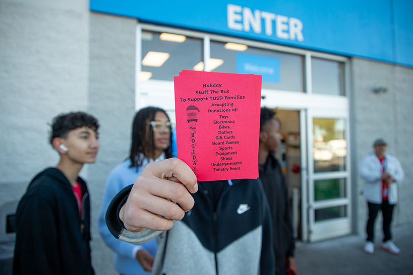 A man holds up a red piece of paper in front of his face with a list of items requested for the Stuff the Bus event