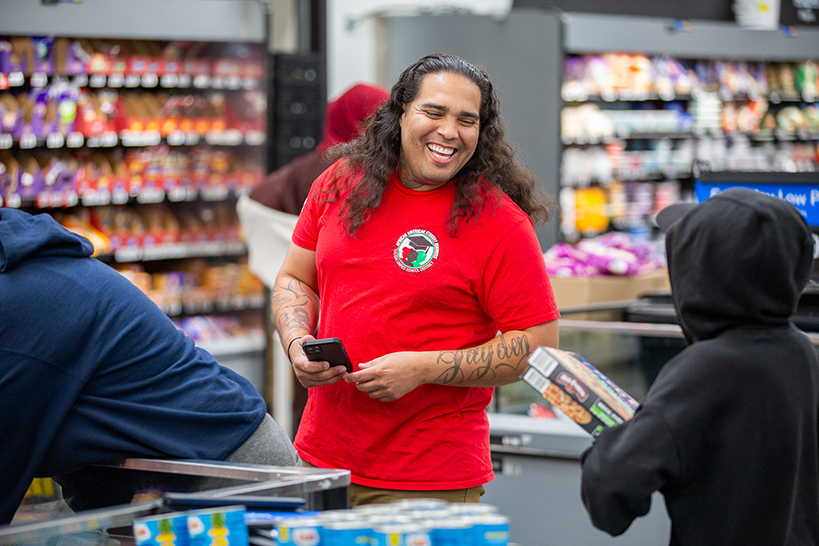A man with curly hair and a red t-shirt laughs as he stands in a checkout line with a teen in a black hoodie