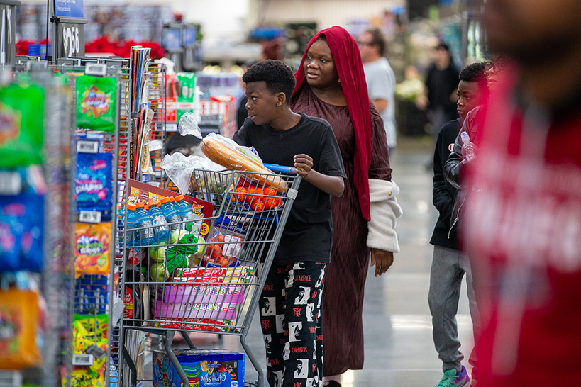 A boy pushes a cart filled with groceries, as his mom and brother walk behind him