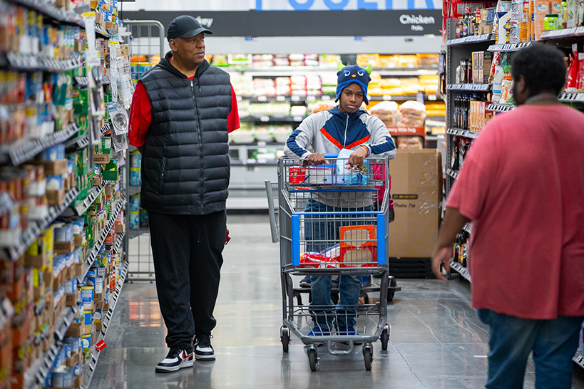 A man walks down a grocery store aisle with his son pushing a cart