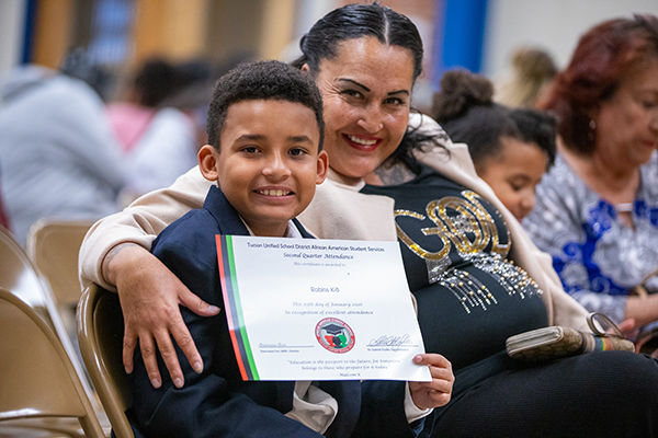 A little boy smiles as he holds his award certificate, with his mom next to him