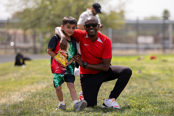 A coach in a red jersey poses next to a young boy in a colorful soccer jersey on the field