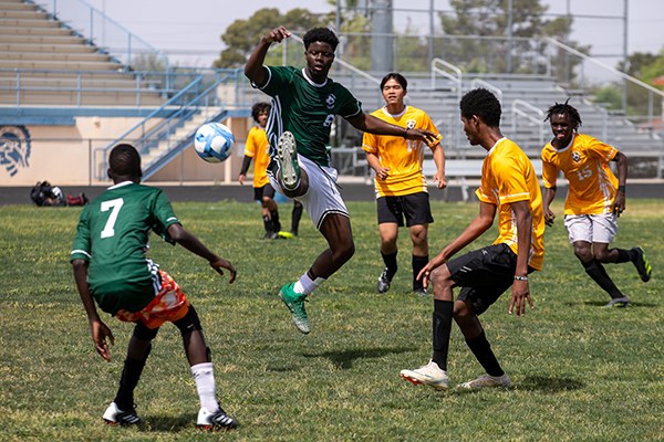 A teen boy in a green soccer jersey kicks the ball as members of the opposing team surround him