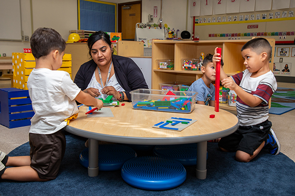 A woman sits on the floor, working with two preschool-age boys as they play with blocks on a table