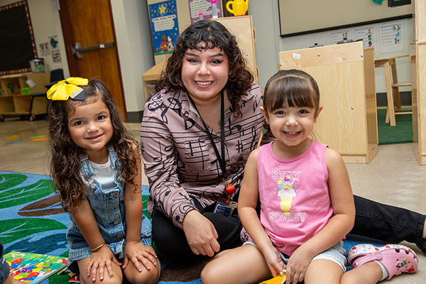 A woman, center, sits with two little girls on a rug