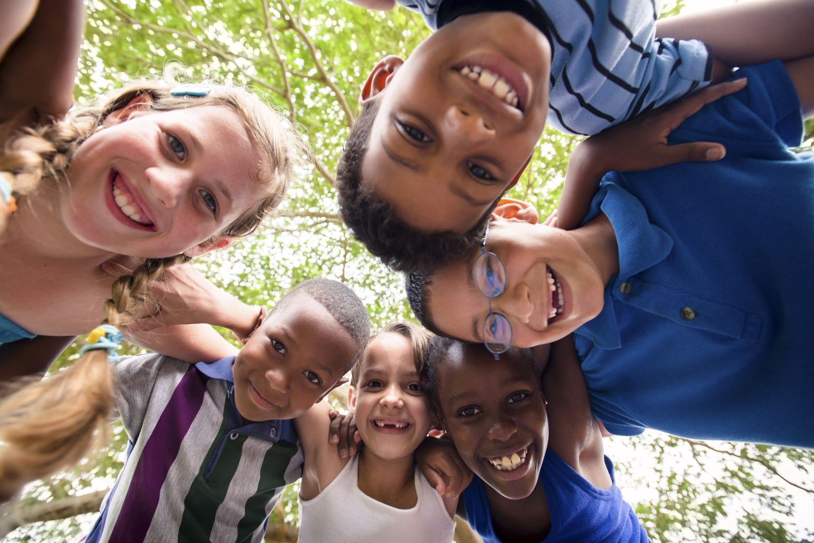 group of 6 racially diverse children circled around and looking down into camera, with arms around one another