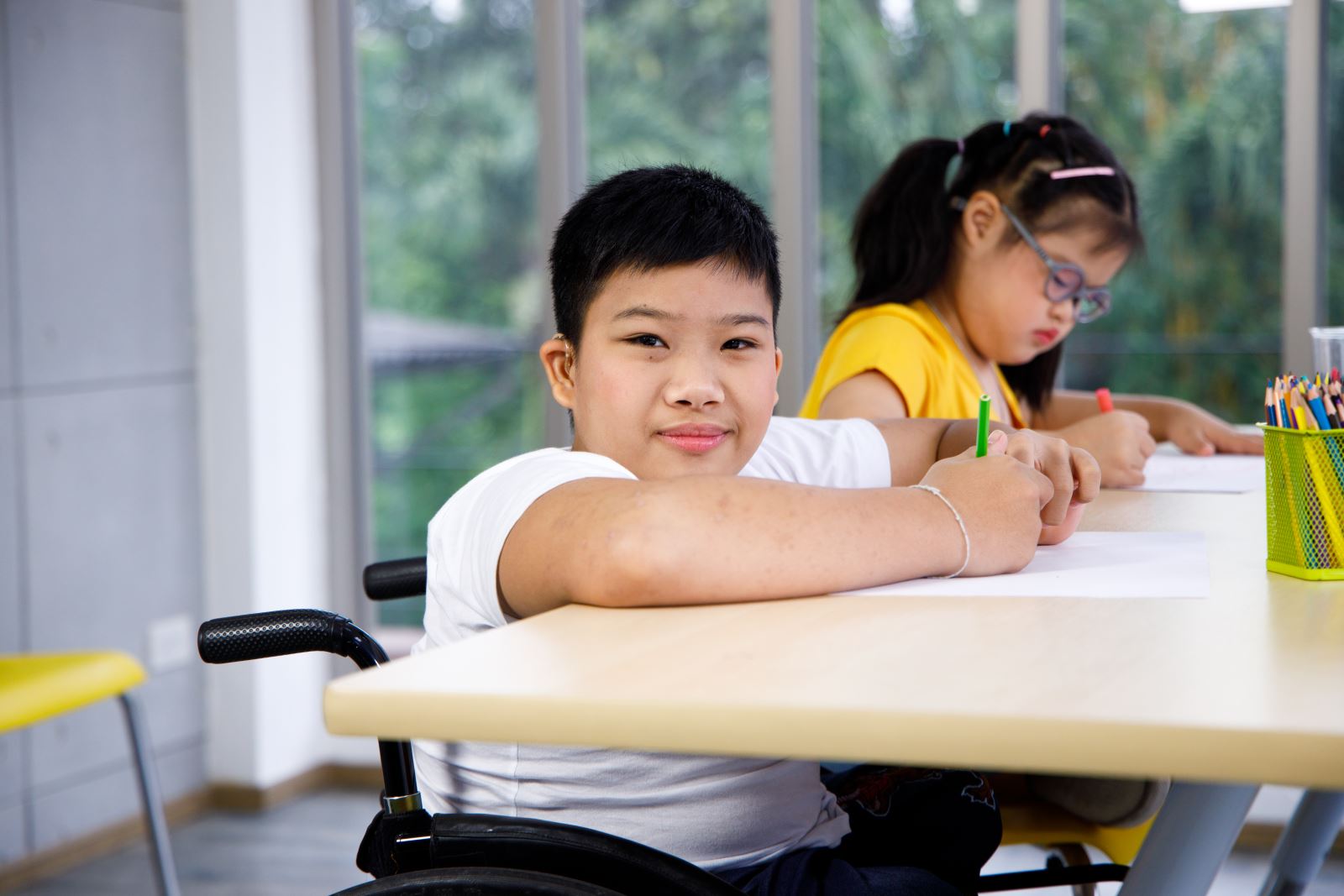 boy in wheelchair with hearing aid and white shirt drawing with girl in yellow shirt with glasses