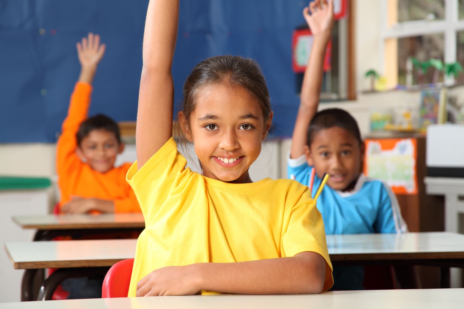 3 non-white smiling preteens in orange, yellow, and blue shirts raise their hands in a classroom