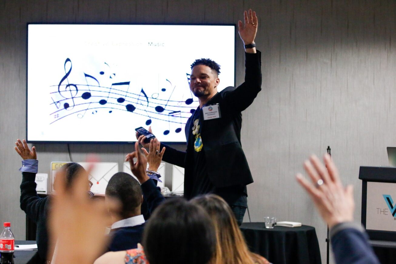 A man in a black suit jacket raises his hand while giving his presentation