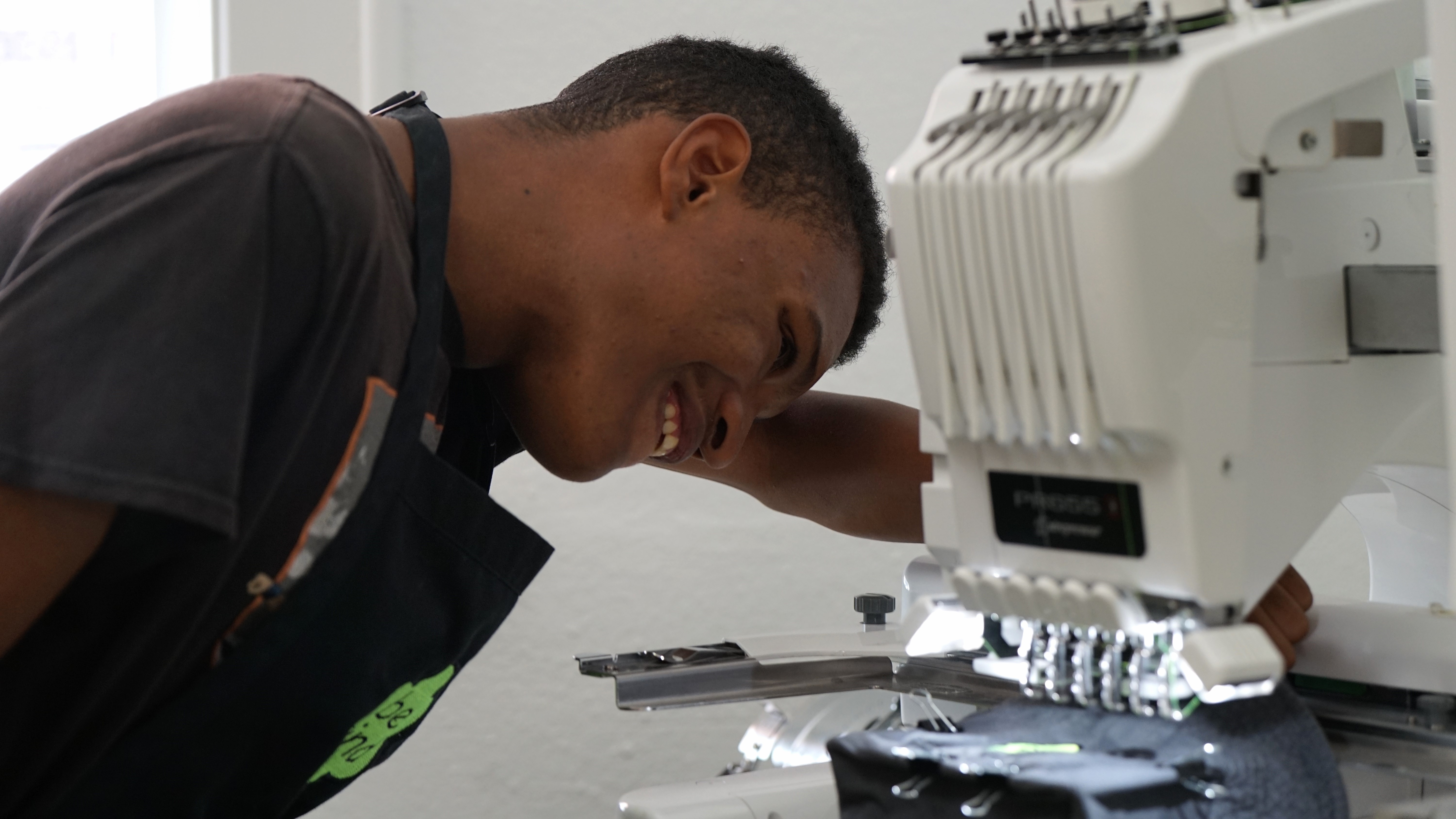 A student smiles as he uses a sewing machine to create a Be Kind logo