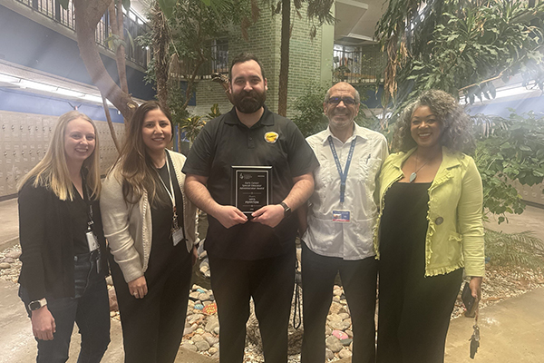 A man, center, holds an award plaque surrounded by 4 colleagues