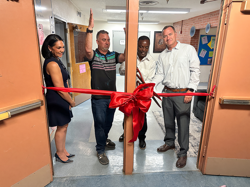 A man cuts a red ribbon with a giant pair of scissors while a woman and two men look on