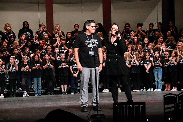 A woman talks into a microphone next to a man, in front of a group of choir students