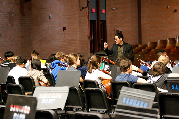 A man wearing glasses and a suit jacket directs a group of student guitarists