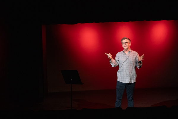 A man in a gray plaid shirt and glasses gives stage directions on stage with red lights