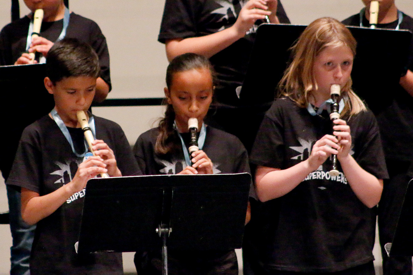 Teacher holds sheet music while a classroom of students listens in the background.