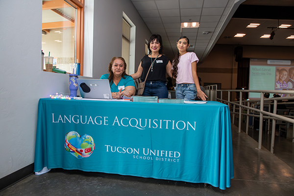 Two women and a preteen girl smile from behind a table with the Language Acquisition tablecloth on it