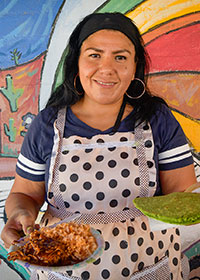 Woman serving traditional food in front of colorful mural.
