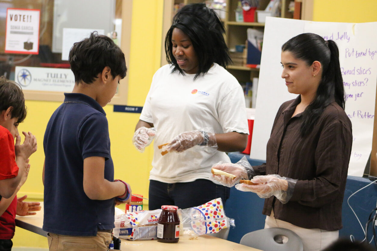 Volunteers help students prepare PB&J sandwiches.