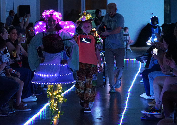 A girl and boy in light-up garden-themed costumes walk the runway