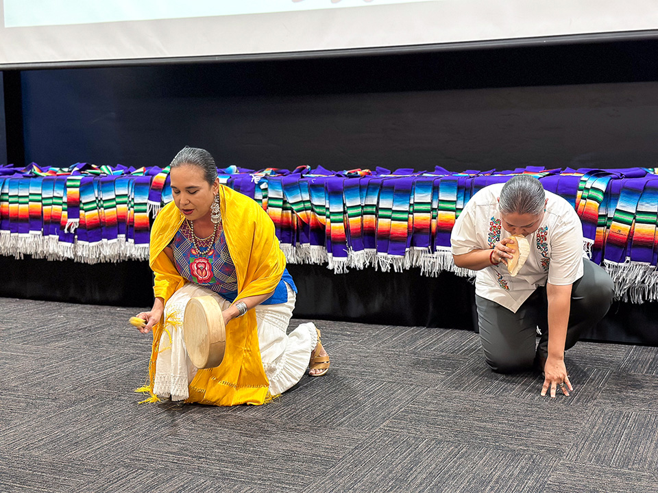 A woman in a yellow scarf kneels on the ground playing a drum, with a man kneeling next to her playing a conch shell