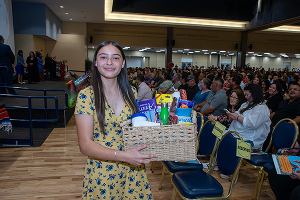 A teen girl wearing a yellow floral dress smiles as she holds up a gift basket