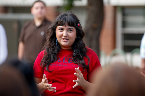 A woman with long dark hair wearing a red shirt addresses a crowd