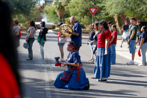 A woman in a blue dress kneels on the ground holding a vessel, while others stand around her