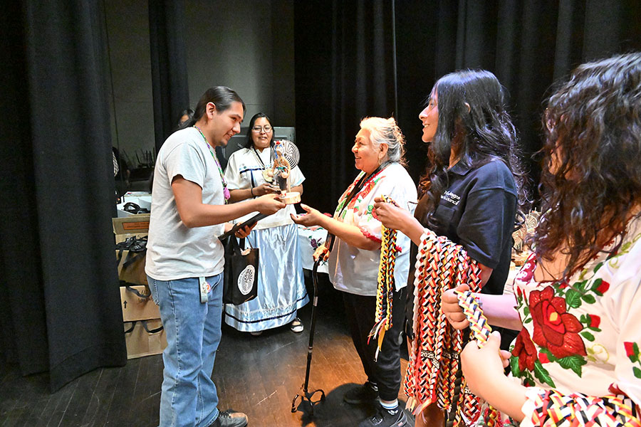 A teen boy holds up his award as a woman gets ready to hand him a woven necklace