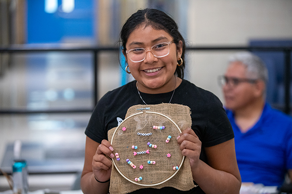A girl wearing glasses smiles as she holds up her beaded creation