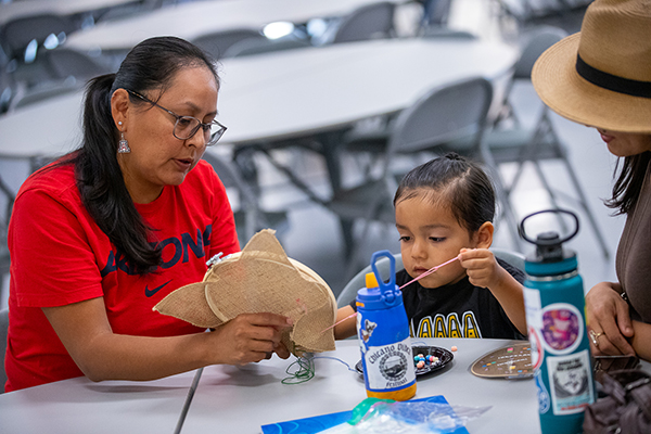 Two women help a little girl as she pulls pink string from a round board