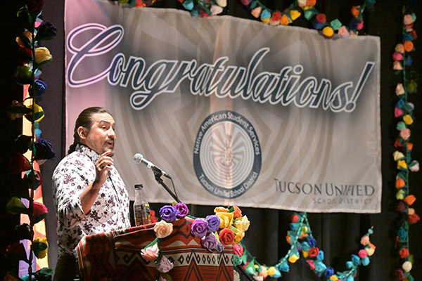 A man talks to the audience on stage with a Congratulations banner behind him