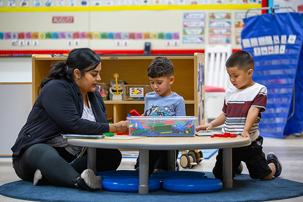 A woman sits on the floor, working with two preschool-age boys as they play with blocks on a table