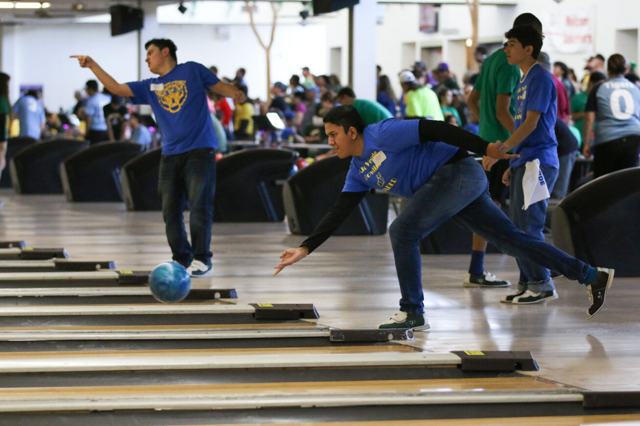 A boy in a blue shirt rolls a blue bowling ball while another boy points at his ball in the next lane.