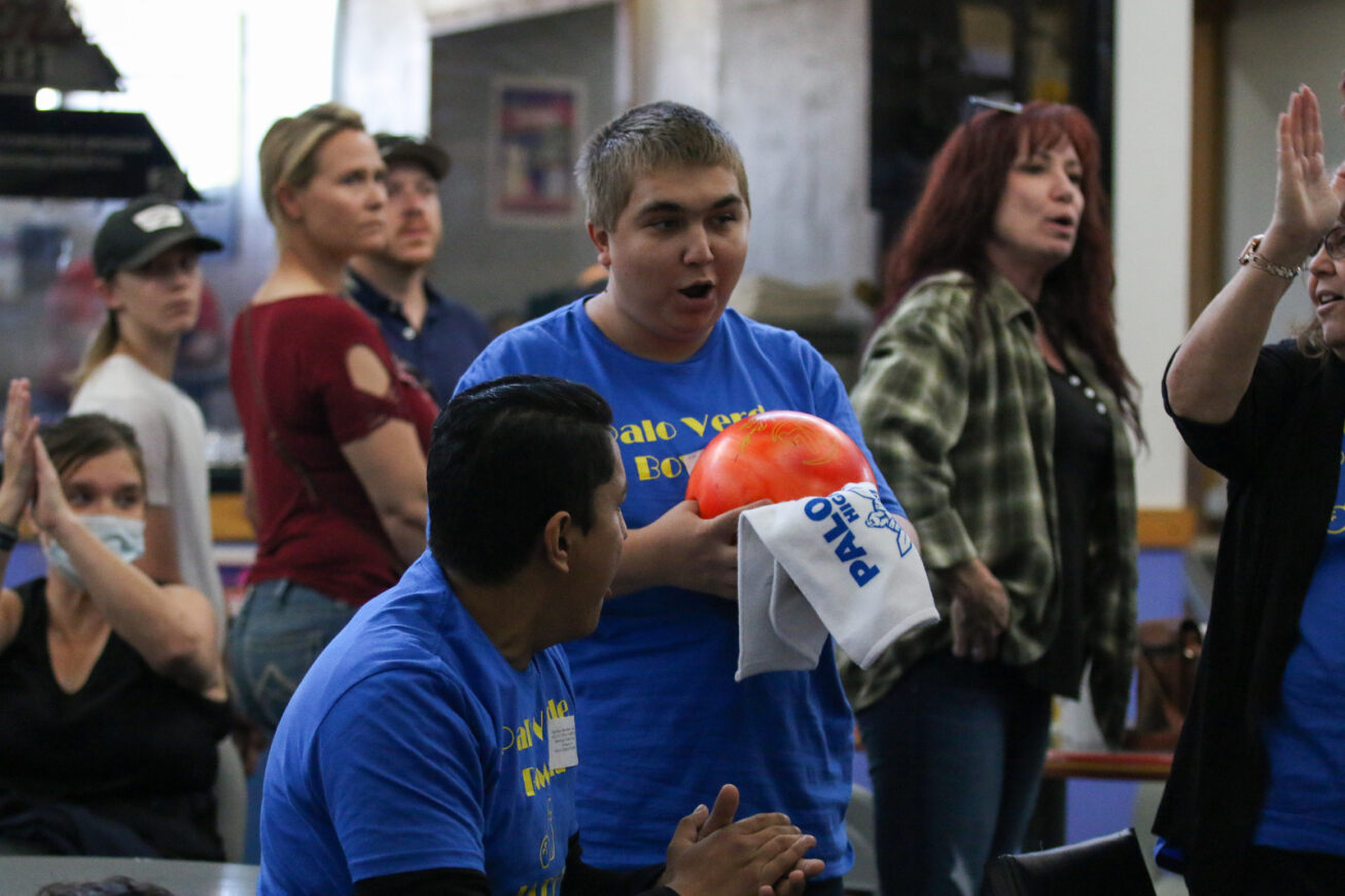 A boy in a blue shirt excitedly shows off his orange bowling ball to a teammate