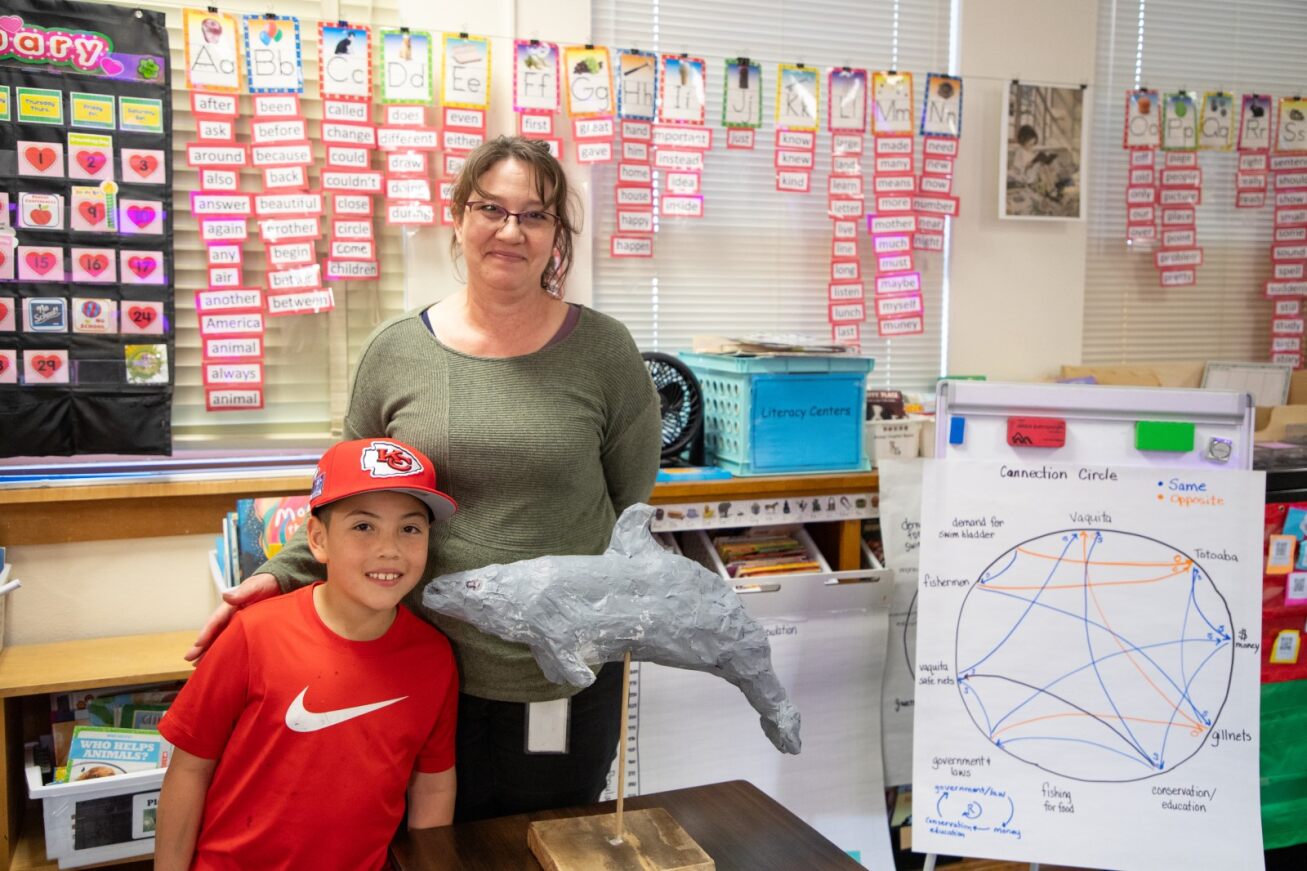 A student in a red hat and t-shirt poses with his 2nd grade teacher and the papier mache vaquita he created