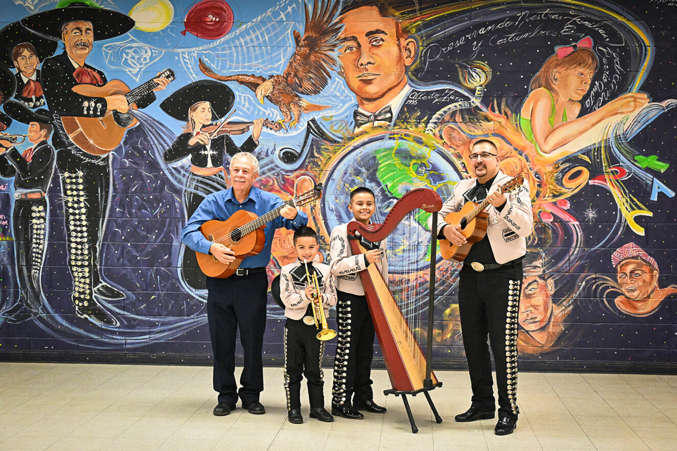 The Valenzuelas hold their instruments in front of a wall decorated with mariachi musicians.