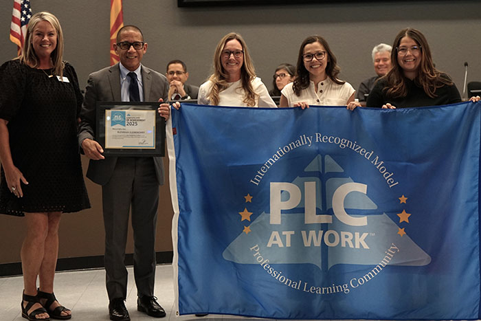 Three women hold up a blue PLC at Work banner while a woman and man next to them hold a certificate